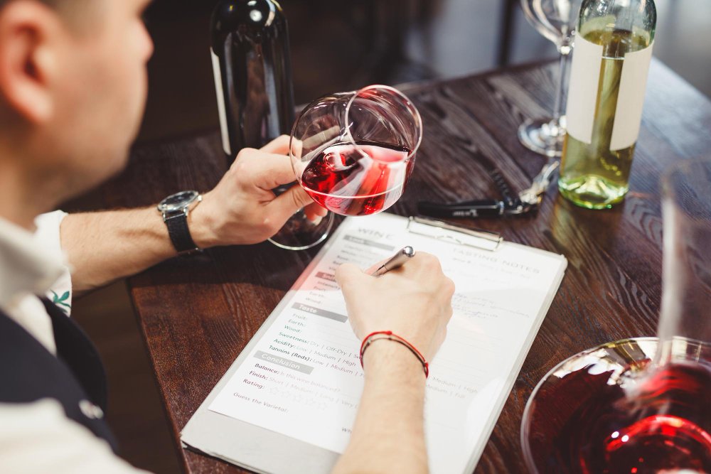 Un homme écrit des notes de dégustation tout en tenant un verre de vin rouge, entouré de bouteilles et de verres.